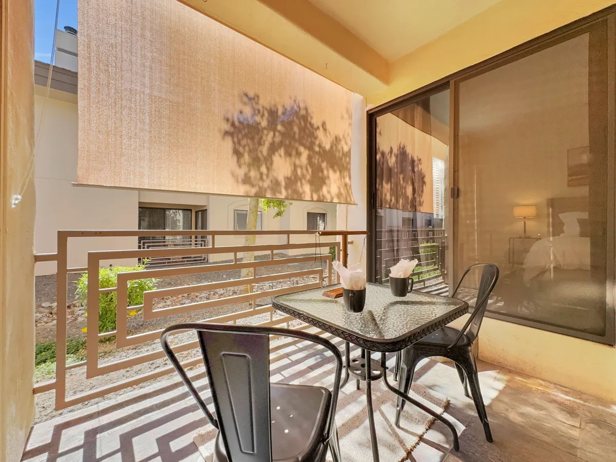 A private patio with a sleek black table and matching chairs, set against decorative railings. Dappled sunlight creates playful patterns, and the space suggests tranquil moments for morning coffee or evening relaxation, with views of the courtyard beyond.