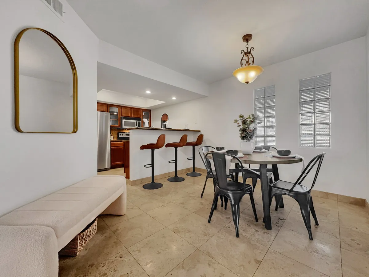 This interior scene features a stylish dining table set with chairs, bar stools lining a kitchen island, a decorative mirror on the wall, and a vibrant flower arrangement. The kitchen and dining areas are open-concept, filled with natural light, and showcase modern finishes throughout. The setup is clearly designed for both casual meals at the breakfast bar and more formal gatherings at the dining table.