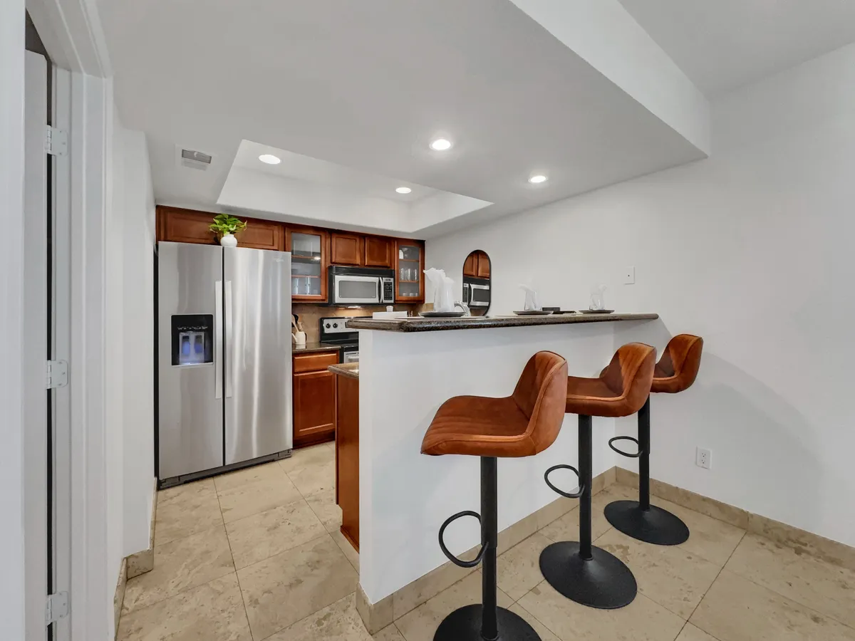 A contemporary kitchen interior featuring a kitchen island with bar stools, stainless steel appliances, and wood cabinetry. The island is set up for casual dining or conversation, highlighting the functionality of the kitchen for both cooking and socializing.