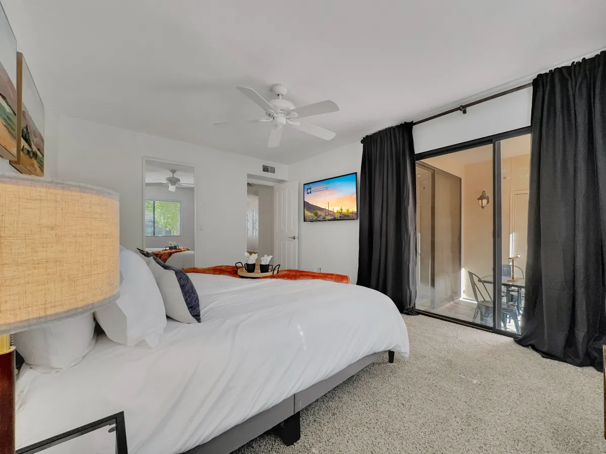 A spacious bedroom with a large bed dressed in white linens, black curtains framing a window, a television mounted on the wall for in-bed viewing, and a lamp providing warm accent lighting. A small dining area is visible through the glass door, which likely leads out to a patio, emphasizing direct outdoor access.