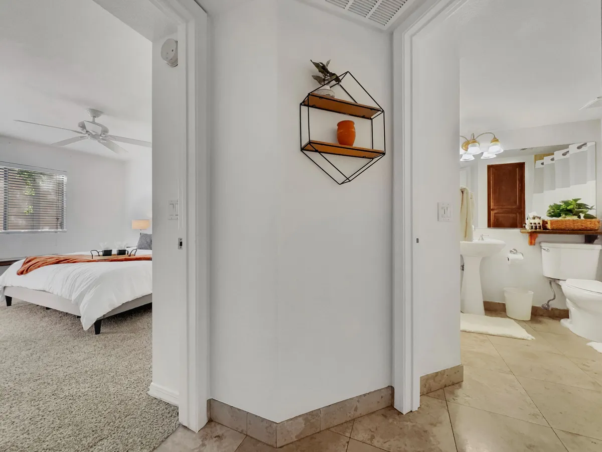 A comfortable bedroom scene featuring a decorative shelf, a view toward the bathroom entrance, and a glimpse of the bed area. The setting is bright and inviting, with natural light filling the room, and a sense of relaxation and readiness for the day.