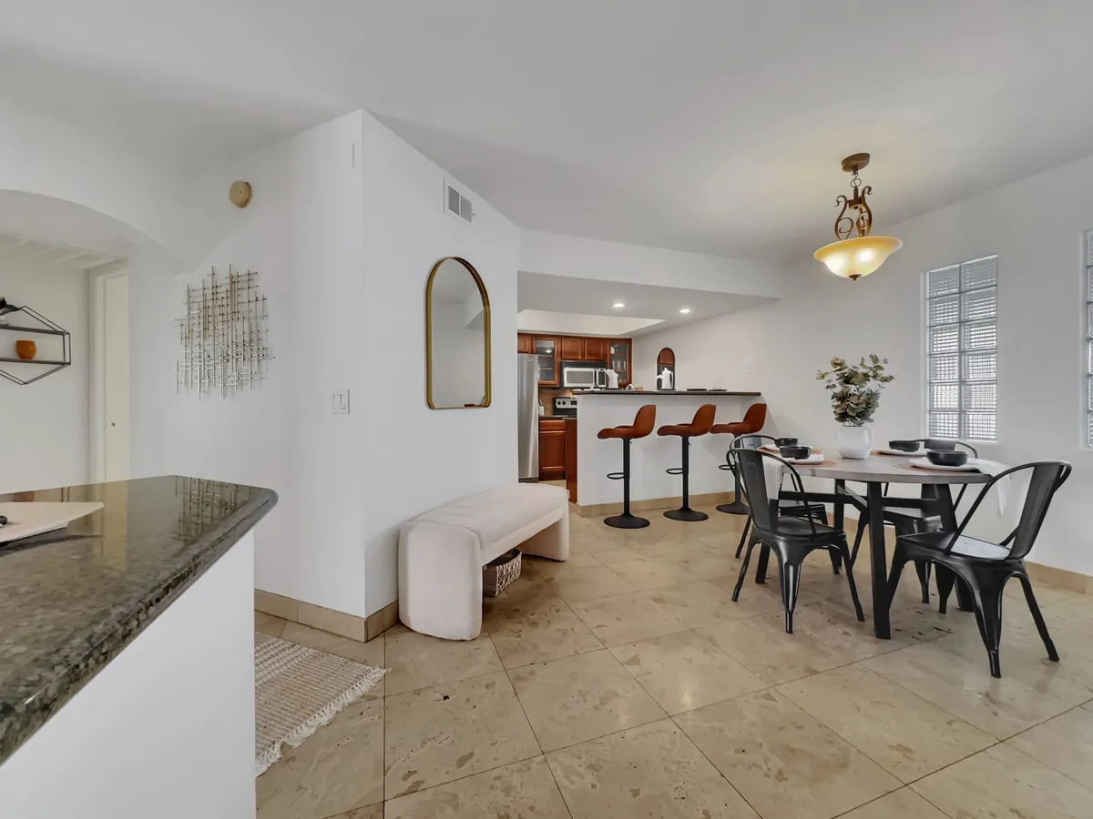 Another inviting view of an open kitchen and dining area. The image shows a dining table with comfortable seating, bar stools by the kitchen island, a decorative plant, and a large mirror. The mood is warm and welcoming, ideal for both family meals and casual gatherings.