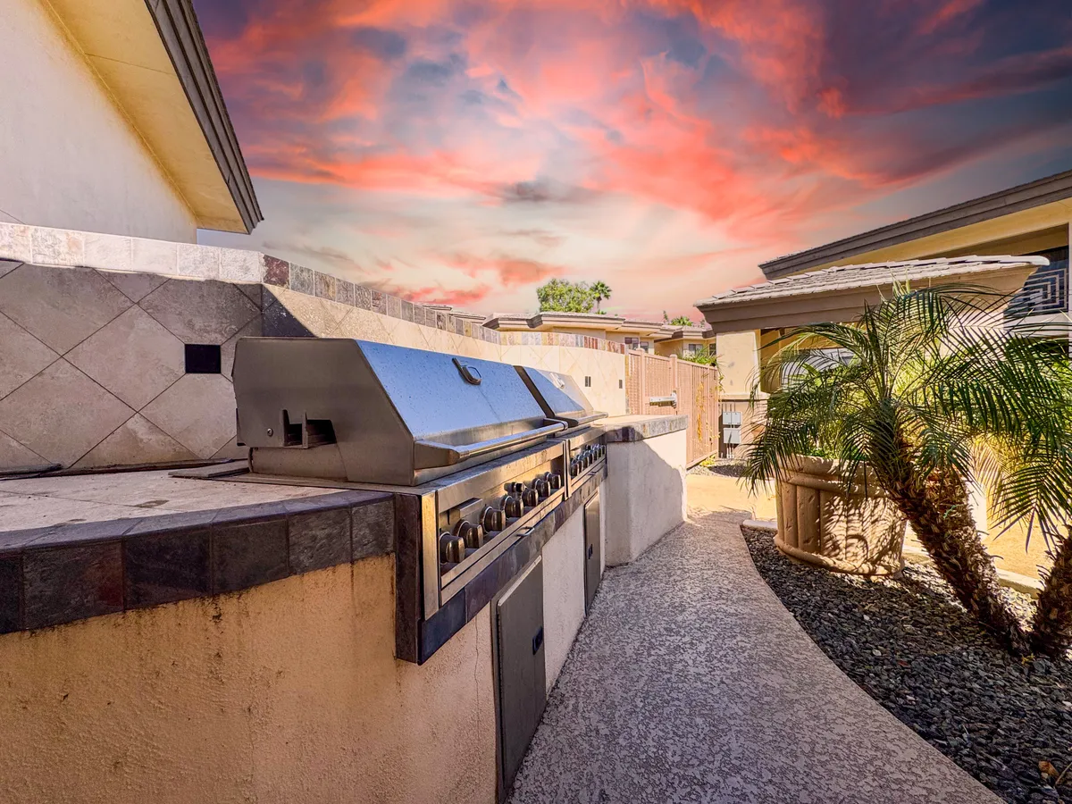 An outdoor grilling area with stone countertops and a palm tree, set against a partly cloudy sky, offers a picturesque spot for barbecuing and gathering with friends or family.