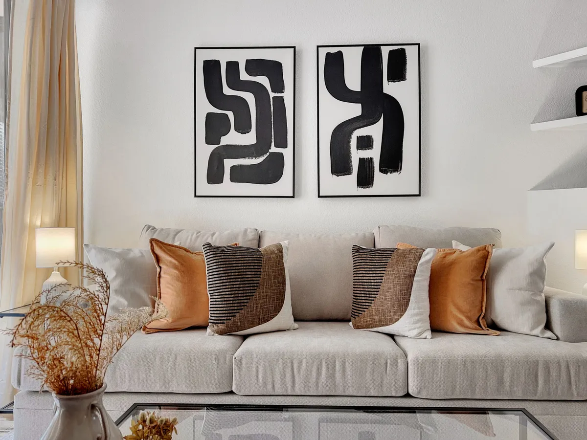 A bright, modern living space featuring a comfortable sofa with decorative pillows, striking abstract wall art, and a glass coffee table. The room invites relaxation and conversation, with an open, airy feel and tasteful decor.
